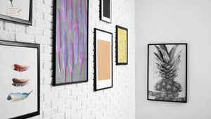 Gallery wall of framed artwork on white brick, with colourful abstract prints, feather illustrations and a black and white pineapple photograph.