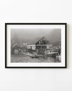 Abandoned British Columbia boathouse and moored boat beneath a misty forest, in black and white.