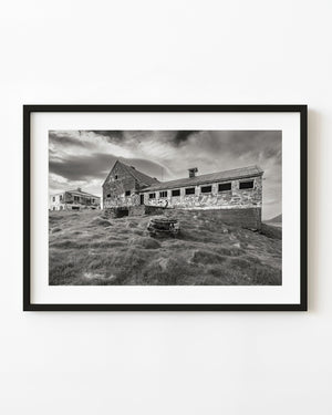 Abandoned Iceland farm buildings with graffiti above a grassy slope, photographed in monochrome.