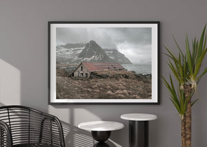 Abandoned coastal boathouse among dry grass beneath a pointed snowy mountain.