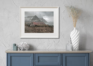 Old Norwegian boathouse beneath cloud-softened mountains on a rugged coast.