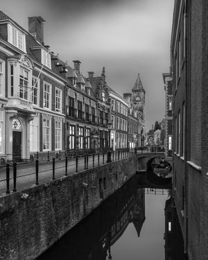 Ein schmaler Kanal führt zu einer Bogenbrücke unter historischen niederländischen Fassaden und einem entfernten Uhrturm.