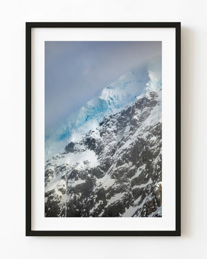 Blue glacier ice above dark rock and snow on an Antarctic mountain beneath low clouds.