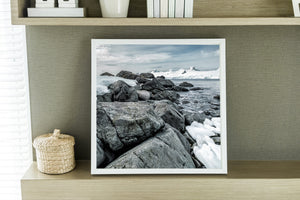 Quadratisches antarktisches Küstenfoto mit Steinen, Meer und fernen Gipfeln.