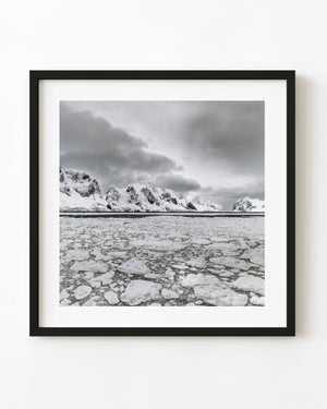Broken Antarctic sea ice beneath snowy mountains and storm clouds.