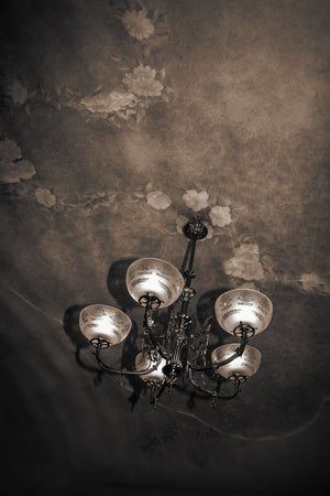 Des abat-jour en verre décoratifs et des ferronneries de lustre incurvées se trouvent sous un plafond floral usé.