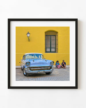 A blue vintage Chevrolet beside seated women against a yellow Havana wall.