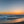 Gentle surf meets a dark shoreline beneath a glowing orange and blue evening sky.
