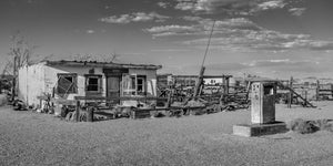 An old roadside fuel pump stands before weathered buildings and fencing on Route 66.