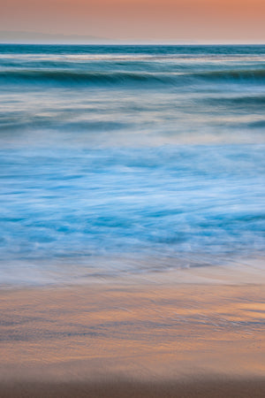 Gentle blue surf meets reflective wet sand in a vertical California coastal photograph.