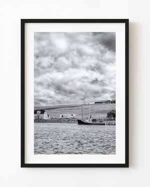 Schwarz-weißes Küstenfoto von Cape Breton mit einem Boot, einem Haus am Hang und strukturierten Wolken.