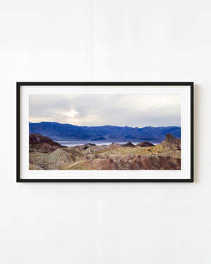 Bunte Death Valley-Kämme erstrecken sich in Richtung entfernter blauer Berge unter einem bewölkten Himmel.
