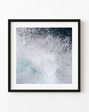 Ocean spray bursts into white foam above pale turquoise water in a square close-up photograph.