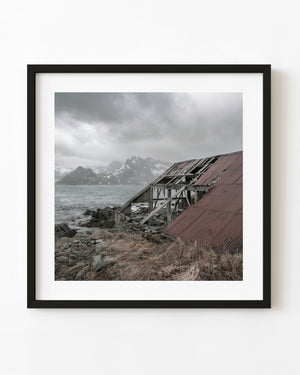 Ein verfallendes Lofoten-Bootshaus mit einem kaputten roten Dach steht neben den schneebedeckten norwegischen Bergen und dem Meer.