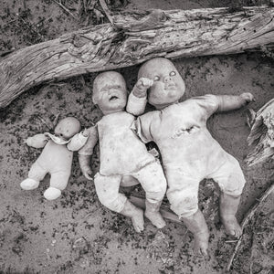 Worn toy dolls lying in sand below a fallen piece of wood, monochrome photograph.