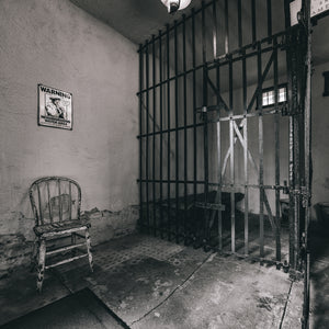 Square monochrome photograph of a historic jail interior with barred doors and cracked plaster
