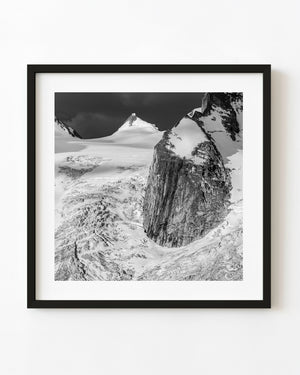 A sheer rock mountain rises beside a crevassed glacier in a black and white photograph