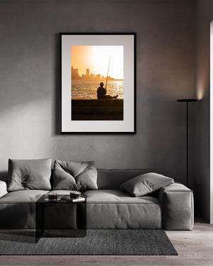 Photographie couleur d'un pêcheur se reposant sur le Malecon alors que le coucher de soleil brille sur l'eau.