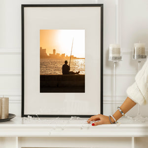 Photographie du coucher de soleil sur le Malecon avec un pêcheur assis, de l'eau scintillante et l'horizon de La Havane.