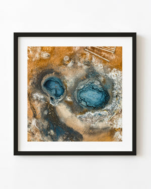 Luftaufnahme von zwei blauen geothermischen Kratern, umgeben von ockerfarbener Erde und hellen Mineralablagerungen in Island.
