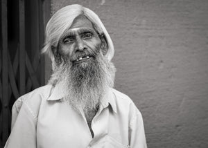 An older Indian man with white hair and a flowing beard looks directly toward the camera.