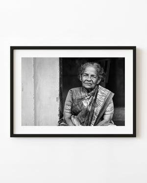 Black and white portrait of an older Indian woman in a patterned saree seated in a dark doorway.