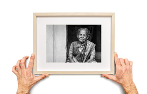 Older Indian woman seated beside a textured white wall, looking slightly away from the camera.
