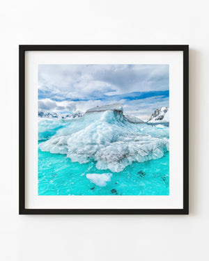 Layered Antarctic iceberg surrounded by bright turquoise water and snowy mountains.