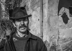 Uomo baffuto che indossa un cappello a tesa scura accanto a un muro esposto alle intemperie