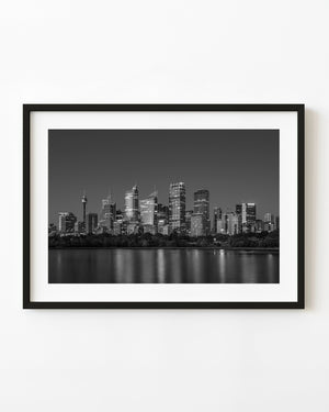 Die Lichter der Skyline von Sydney spiegeln sich in einem Schwarz-Weiß-Nachtfoto im glatten Hafenwasser.