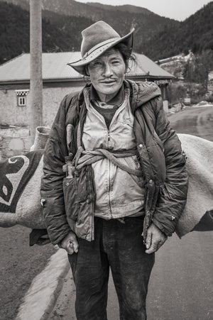 A man faces the camera beside a mountain road with a cloth bundle behind his shoulders.