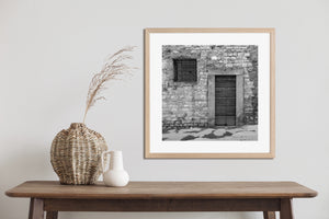 Fotografía monocromática cuadrada de mampostería rústica, una puerta de madera y pavimento húmedo.