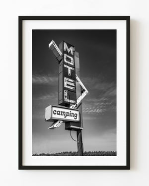 Vintage motel and camping sign beneath a cloud-streaked sky in black and white.