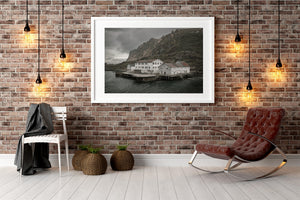 Weathered coastal buildings above the water beneath rugged Norwegian cliffs