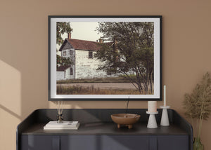 Casa de campo blanca con pintura desgastada y chimeneas de ladrillo enmarcadas por ramas frondosas