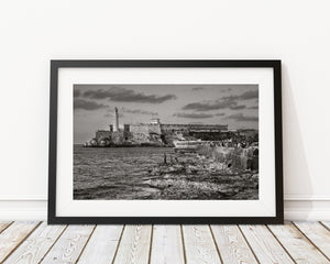 Figures on coastal rocks beneath a curved Havana seawall with Morro Castle in the distance.