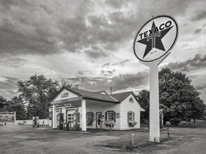 A round Texaco emblem rises above a small white gas station and vintage pumps under textured clouds.