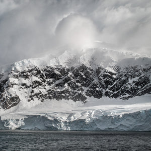 Dunkler Fels, blaues Eis und Schnee entlang der antarktischen Küste unter blassen Wolken.