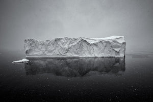 Falling snow surrounds a pale Antarctic iceberg above its dark reflection.