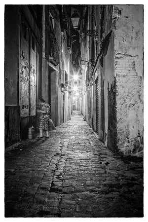 A stone-paved Gothic Quarter passage recedes past a woman at a dark doorway in black and white.