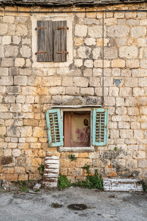 Una fotografía vertical en color muestra dos ventanas en una pared de piedra pálida con contraventanas azul verdosas debajo.