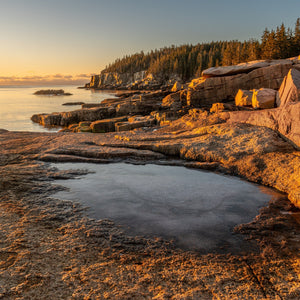 Sunrise warms Acadia’s rocky shoreline beside a frozen foreground puddle and calm sea