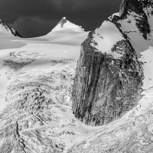 Snow and glacier ice surround a steep exposed rock face beneath a dark sky