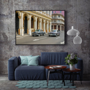 Classic Cuban cars drive past ornate doors and a yellow-and-white colonnade.